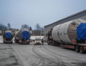 Drei große Edelstahlsilos auf Tiefladern, abgedeckt mit blauen Planen, bereit zum Abtransport auf einem verschneiten Betriebsgelände mit Lastwagen und Pkw.