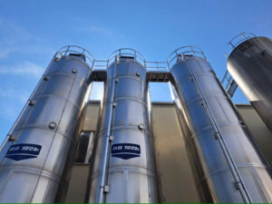 Drei große, glänzende Edelstahlsilos von RB Tech vor blauem Himmel, angebracht an einer hellen Gebäudeaußenwand, mit Leitern und Geländern auf der Oberseite