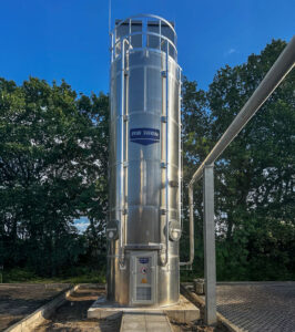 Hohes, zylindrisches Außensilo aus Edelstahl von RB Tech mit zwei Kammern, stehend auf einem Fundament, umgeben von Bäumen und blauem Himmel.